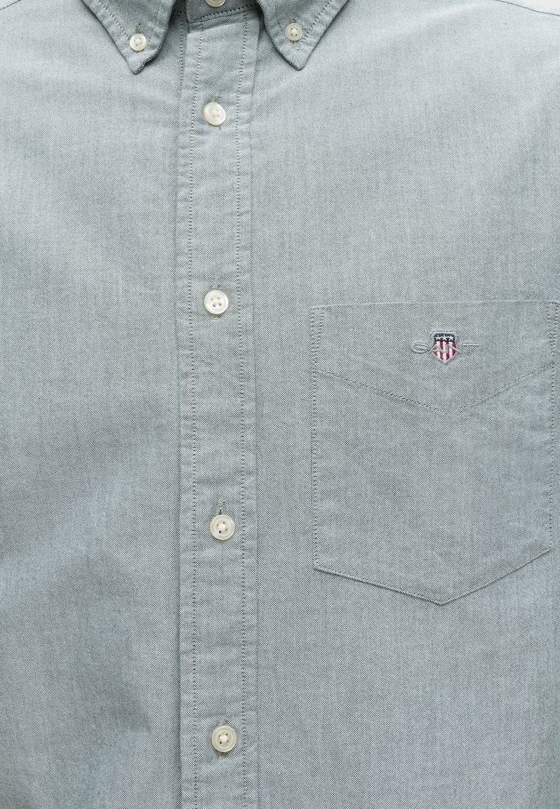 Camisa gris claro abotonada con un bolsillo en el pecho que presenta un pequeño emblema bordado en forma de escudo con estrellas y franjas.