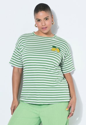 Frau mit kurzen Haaren, die ein grün-weiß gestreiftes Shirt mit Zitronenaufnäher und hellgrüne Hose trägt, steht vor einem weißen Hintergrund.