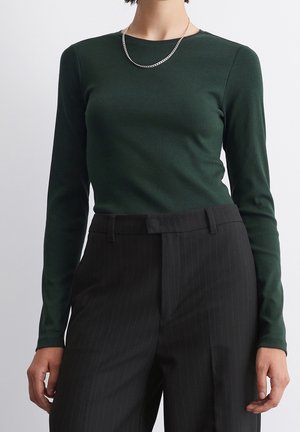 Person trägt ein dunkelgrünes Langarmoberteil, schwarze Nadelstreifenhose und eine silberne Kette vor einfarbigem Hintergrund.
