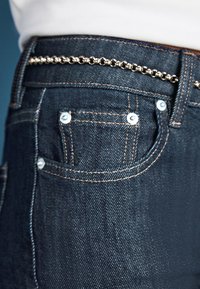 Tasca dei jeans in denim blu scuro con cuciture visibili e rivetti metallici, accessoriata con una sottile cintura a catena d'oro infilata nei passanti della vita.