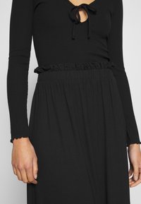 Top noir à manches longues avec un col en V, doté d'un détail noué au niveau du col et d'un ourlet volanté. Le tissu semble doux et extensible.
