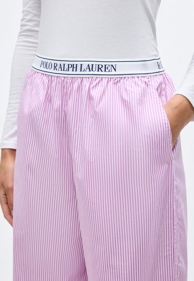 Άτομο που φοράει ριγωτό παντελόνι σε ροζ και λευκές κάθετες ρίγες με λευκή ζώνη μέσης που αναγράφει "POLO RALPH LAUREN" και λευκή μπλούζα με μακριά μανίκια.