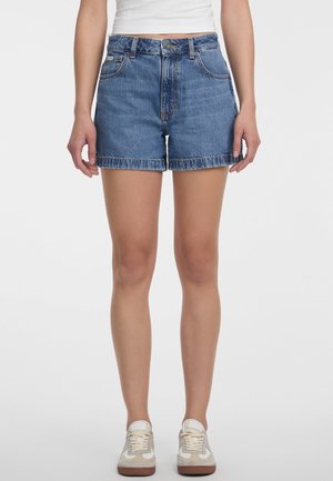 Kvinde iført blå denimshorts, hvid cropped top og hvide sneakers, stående mod en ensfarvet baggrund med hænderne afslappet ned langs siderne.