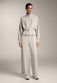 Vaaleanharmaa aktiivivaatetussetti, joka sisältää vetoketjullisen, korkeakauluksisen collegepaidat, jossa on peplum-vyötärö, sekä leveät, laskostetut housut. Valmistettu pehmeästä kankaasta.