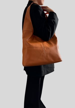 Una bolsa tote de cuero marrón con una textura suave, que presenta costuras lisas y un diseño espacioso, sostenida por una persona con un atuendo negro.