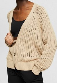 Beige gestrickte Strickjacke mit lockerem Sitz, V-Ausschnitt, vertikalen Streifen und Knöpfen auf der Vorderseite. Strukturierte offene Webart verleiht optisches Interesse.
