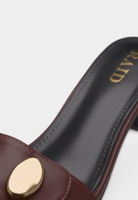 Mule en cuir marron avec semelle intérieure noire et décoration ovale dorée sur la sangle supérieure.