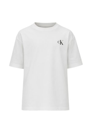 Giovane ragazzo con capelli ricci che indossa una t-shirt bianca Calvin Klein, pantaloni neri a gamba larga e scarpe nere, in piedi su sfondo bianco.