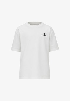 Giovane ragazzo con capelli ricci che indossa una t-shirt bianca Calvin Klein, pantaloni neri a gamba larga e scarpe nere, in piedi su sfondo bianco.