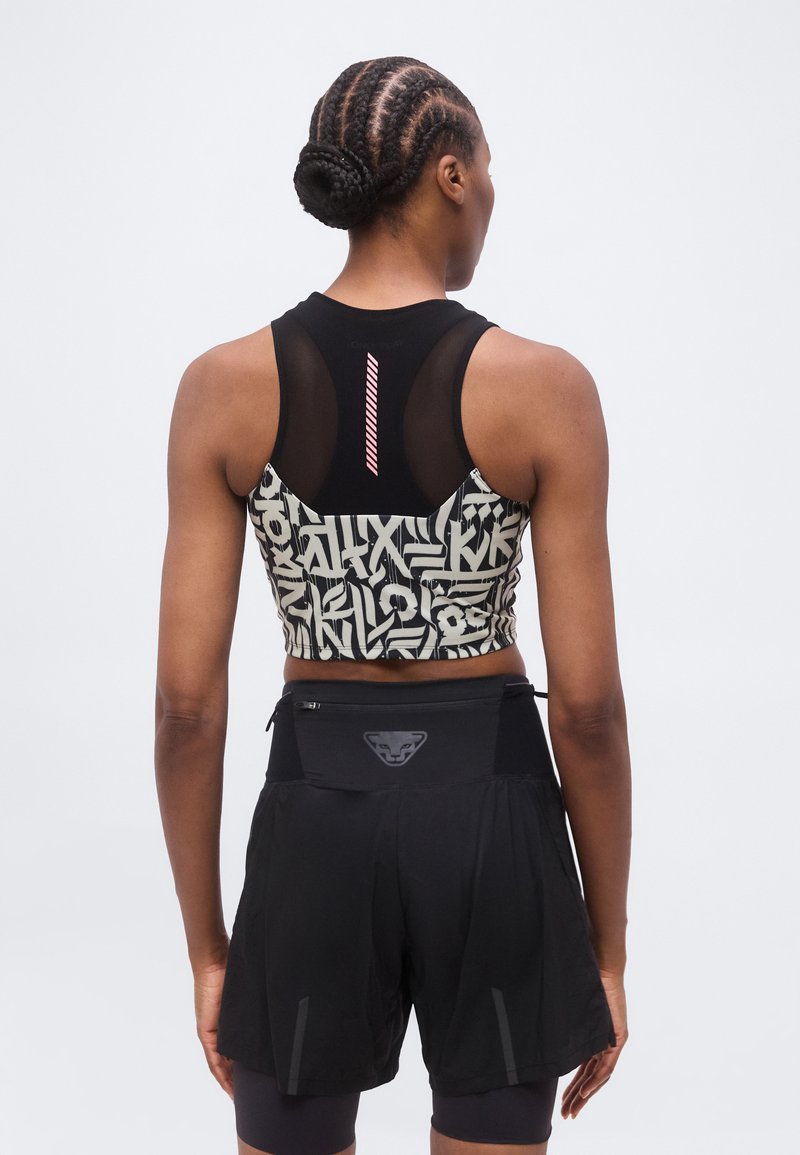 Γυναίκα με πλεξούδες στα μαλλιά, φορώντας αθλητικό τοπ με μαύρο και άσπρο μοτίβο και μαύρο αθλητικό σορτς με τσέπη στη μέση, στέκεται γυρισμένη με την πλάτη.