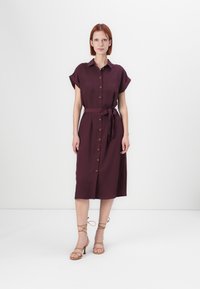 Burgundy kortärmad klänning med knäppning fram, bältesmidja och två bröstfickor. Slät tyg och knälång design.