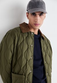 Olive green quilted jacket with brown corduroy collar, snap buttons, and side pocket, worn over a navy shirt, paired with a gray cap.