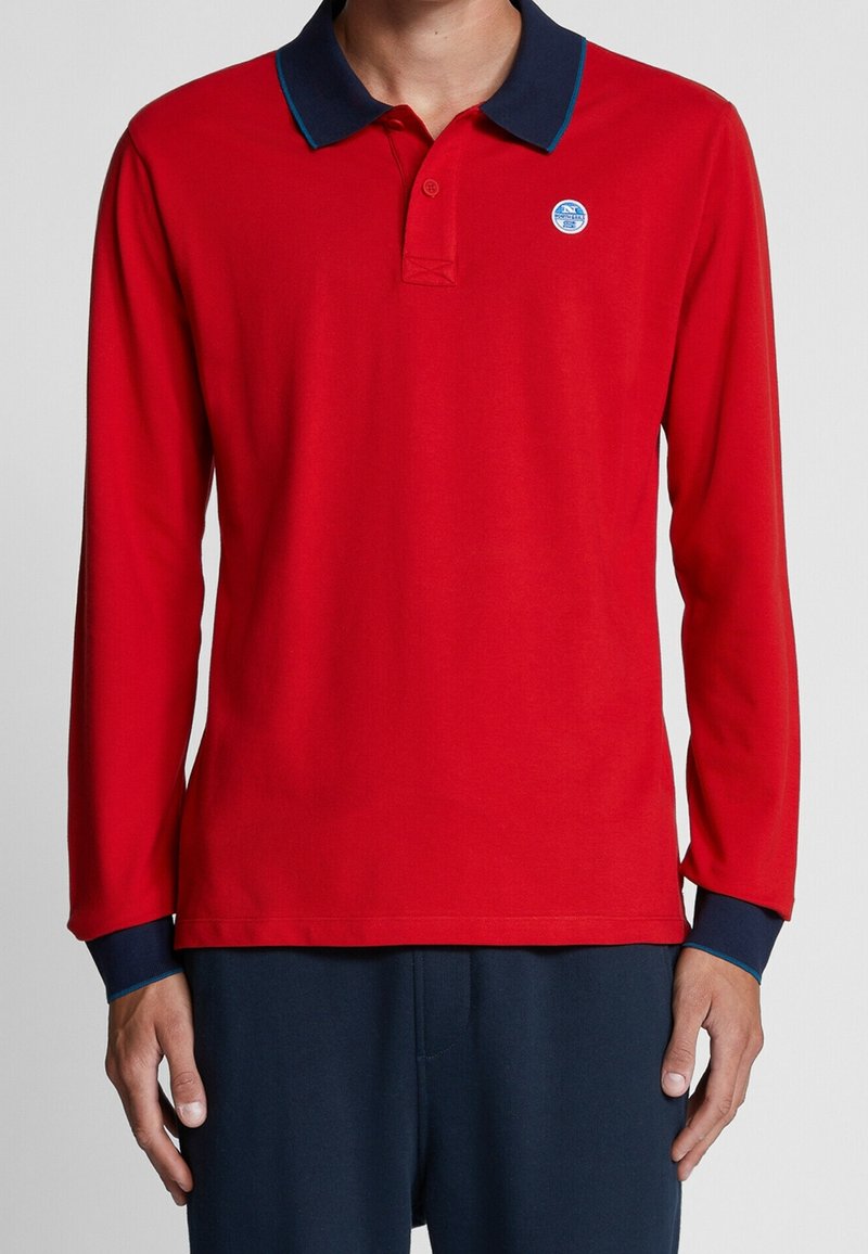 Homme portant un polo rouge à manches longues avec col et poignets bleu marine, et un pantalon bleu marine, debout, les bras détendus le long du corps.