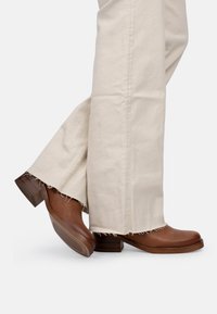Bottines en cuir marron avec un talon carré et une semelle texturée, accompagnées d'un pantalon en crêpe large crème avec les ourlets effilochés.