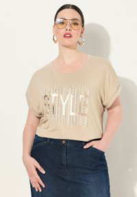Béžové tričko s krátkým rukávem s nápisem "STYLE" tištěným v metalických akcentů a texturovaným designem. Kombinováno s tmavými džínsovými sukněmi.