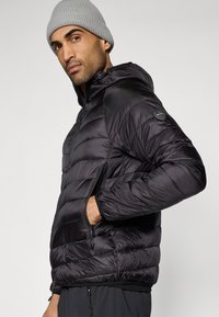 Casaco acolchoado preto isolado com capuz, design acolchoado horizontal, bolsos frontais com zípree, e patch de logotipo no ombro. Combinado com um gorro cinzento.