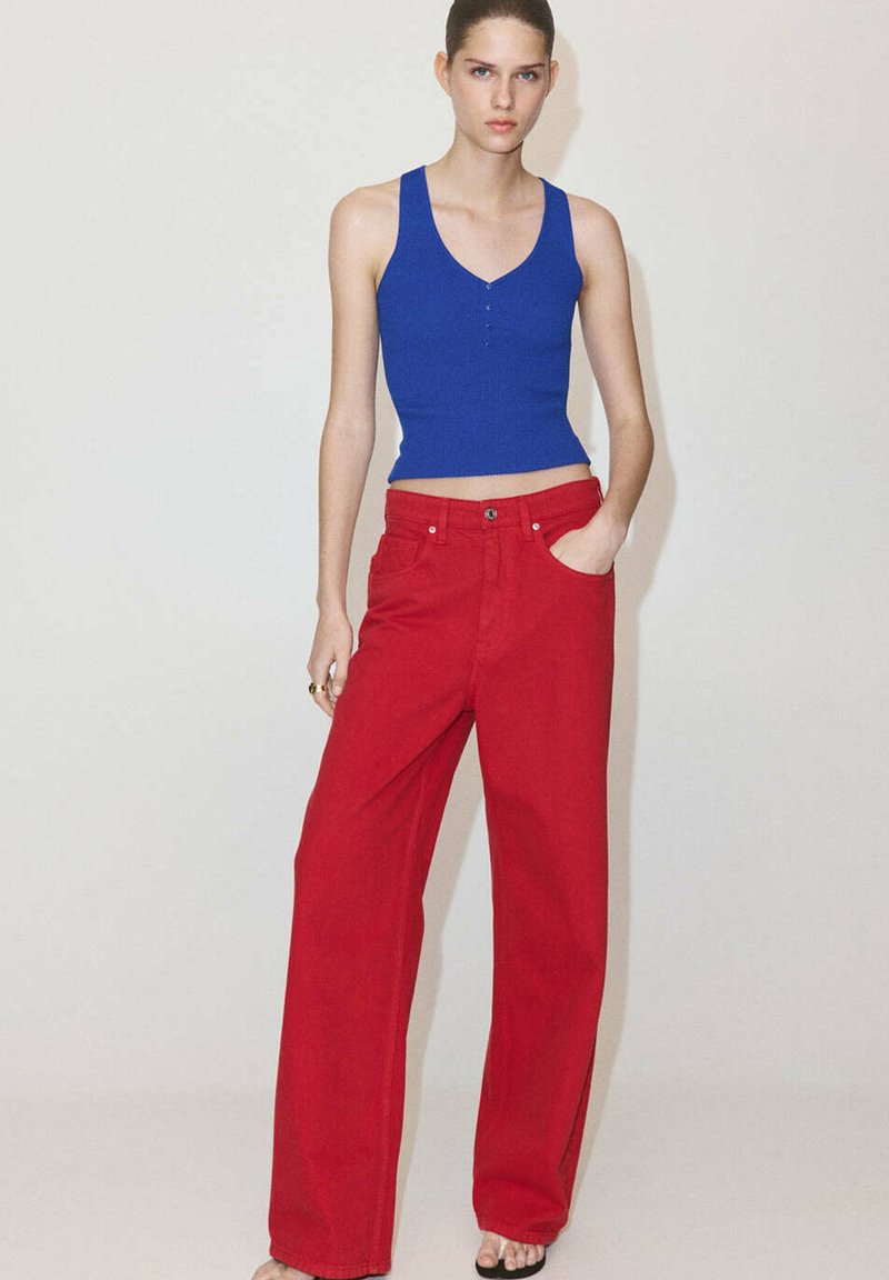 Femme portant un haut bleu sans manches et un pantalon rouge large taille haute, debout devant un fond clair uni.