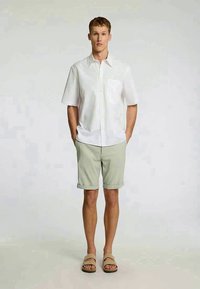 Camisa de botão de mangas curtas branca, calções verde claro e sandálias bege; ajuste casual com um bolso na camisa e as bainhas dos calções arregaçadas.