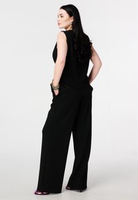 Zwart mouwloos topje gecombineerd met zwarte broek met wijde pijpen, voorzien van gestructureerde stof en zakken. Model draagt paarse schoenen met hak.