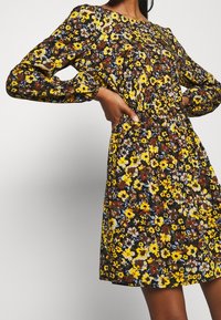 Robe fleurie à manches longues, avec un fond noir orné de fleurs jaunes, bleues et marron. Cintrée à la taille, tissu texturé.