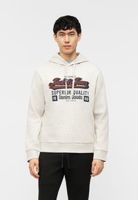 Νέος άνδρας που φοράει ανοιχτό γκρι φούτερ με κουκούλα με την επιγραφή «Jack & Jones Superior Quality Denim Goods 1990» και μαύρο παντελόνι, στέκεται μπροστά σε λευκό φόντο.