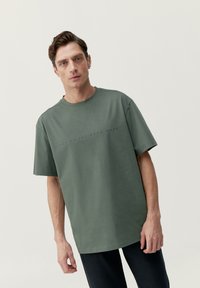 Tričko s krátkym rukávom v tlmenej zelenej farbe z bavlny, s jemnou textovou grafikou "HOP ON THE BORN WAVE" cez hrudník. Voľný strih.