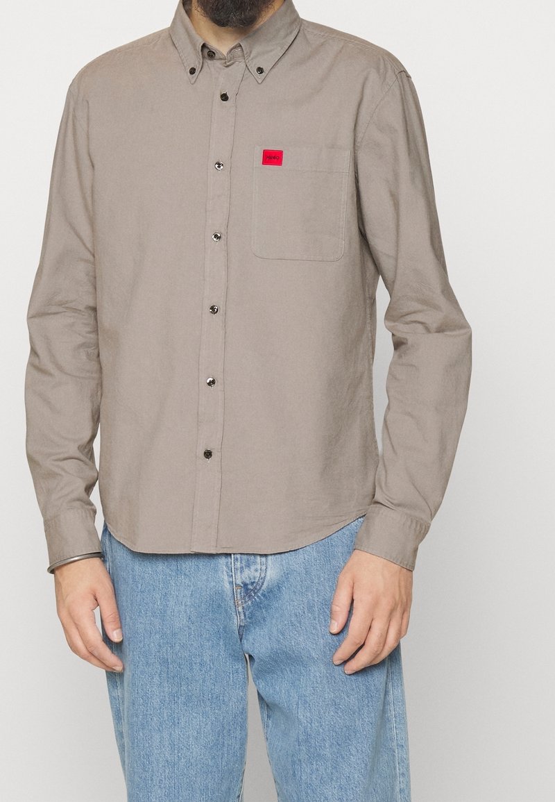 Homme portant une chemise beige boutonnée avec une poche poitrine et une petite étiquette rouge, associée à un jean bleu clair.