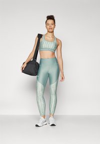 Mujer de pie sosteniendo una mochila de gimnasio negra sobre el hombro, vestida con un sujetador deportivo de patrón verde, leggings a juego y zapatillas de correr blancas.