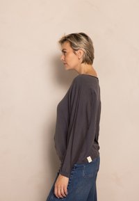 Graues, locker sitzendes Langarmshirt mit tiefem Rundhalsausschnitt, strukturiertem Stoff und einem kleinen Stoffetikett an der Seite, kombiniert mit hoch taillierten blauen Jeans.
