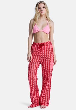 Młoda kobieta z długimi blond włosami, ubrana w jasnoróżowy top bikini i czerwone, wzorzyste, szerokie spodnie, stojąca boso na białym tle.