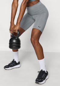 Γκρι σορτς ψηλόμεσα από ελαστικό υλικό, συνδυασμένα με μαύρα και λευκά αθλητικά παπούτσια και κρατώντας μια μαύρη κάτμπελ. Λευκές αθλητικές κάλτσες.