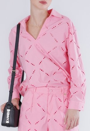 Mujer con blusa rosa con patrón de ojales y pantalones a juego, sosteniendo un bolso de cuero negro con correa para el hombro.