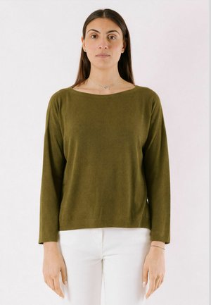 Donna con capelli lunghi e scuri, che indossa un maglione verde oliva a maniche lunghe e pantaloni bianchi, in piedi davanti a uno sfondo chiaro e semplice.