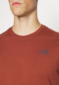 Man som bär en rostfärgad t-shirt från The North Face, visar halsringning, nedre delen av ansiktet och logotypen på övre vänstra bröstet.
