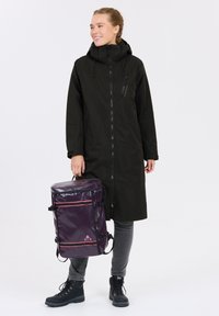 Black long coat with a high collar and zipper, paired with slate gray jeans and black boots. Holding a round purple backpack with red accents.
