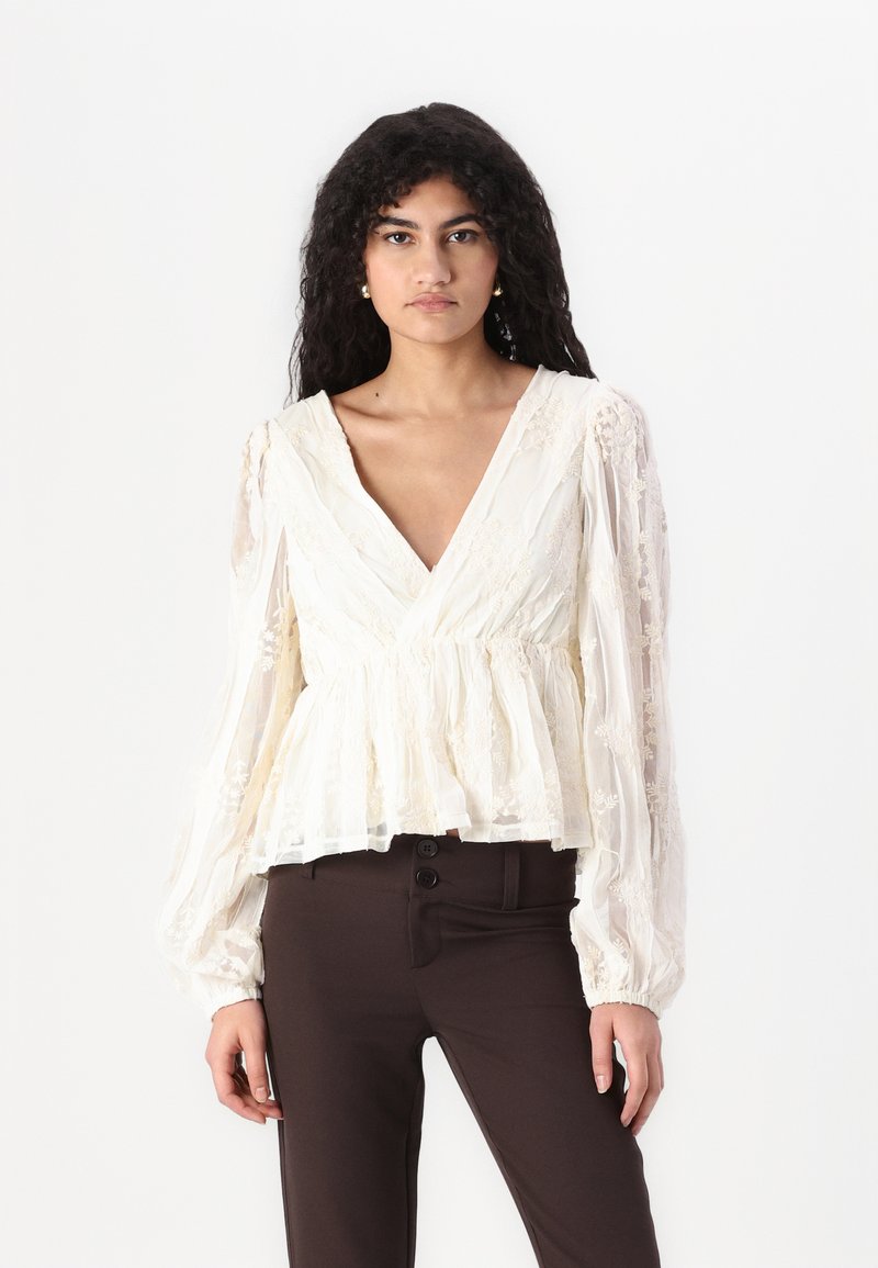 Hvit bluse i blonder med dyp V-hals, lange flagrende ermer, og peplumliv. Strukturert stoff med blomsterdesign og gjennomsiktig finish.