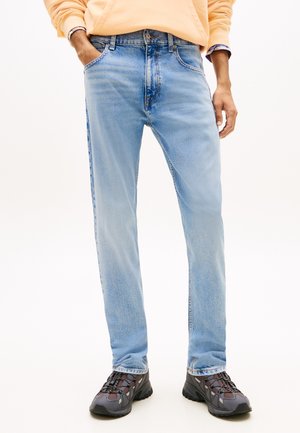 Mand iført lyseblå straight-fit jeans, beige sweatshirt og sorte og grå vandresko, stående med hænderne i lommerne.