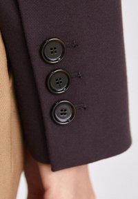 Tela de color púrpura oscuro con una textura suave y tres botones negros brillantes en el puño; los botones tienen cuatro orificios para abrochar.