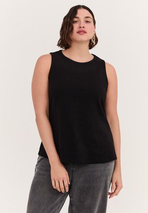 Femme aux cheveux courts et foncés portant des boucles d’oreilles créoles en or, un haut noir sans manches et un jean gris, debout devant un fond uni.