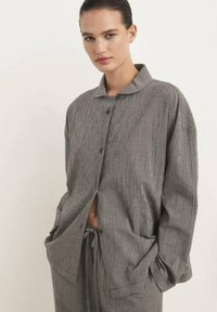 Camicia oversize a scacchi bianco e nero con colletto, bottoni frontali e tasche laterali. Il tessuto appare texturizzato e morbido.