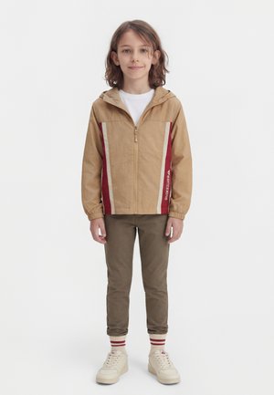 Dítě s hnědými vlasy po ramena, které má na sobě béžovo-červenou bundu na zip, hnědé kalhoty a bílé tenisky s pruhovanými ponožkami.