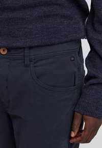 Dark navy pants made of cotton, featuring a front pocket, contrast stitching, and a wooden button at the waist. Soft, textured finish.