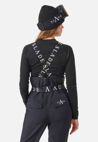 Zwarte lange mouwen top met geribbelde textuur, gelaagd onder bretels met "SLADE" logo. Zwarte broek met zakken en logo detail.