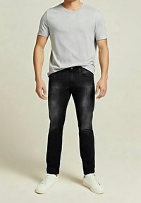 Homme portant un t-shirt à manches courtes gris clair, un jean noir et des baskets blanches, debout devant un fond beige uni.