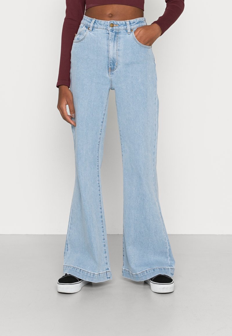 Personne portant un jean large taille haute bleu clair, un top court bordeaux à manches longues, et des baskets noires et blanches, debout avec une main dans la poche.