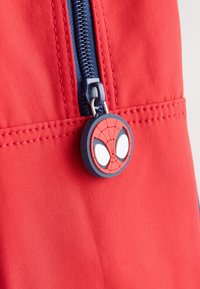 Red fabric with visible stitching, featuring a blue zipper pull adorned with a circular Spiderman logo in red and white.