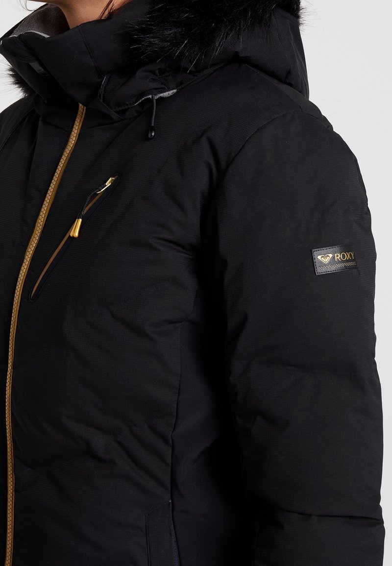 Svart pufferjacka med huva i fuskpäls, gyllene dragkedjor och Roxy-logotyp på ärmen. Strukturerad textil med vanlig passform.