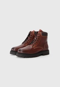 Botas de cuero marrón hasta el tobillo con suelas de goma, punteras redondas y ocho ojetes. Presentan acentos en marrón oscuro contrastantes y costuras visibles.