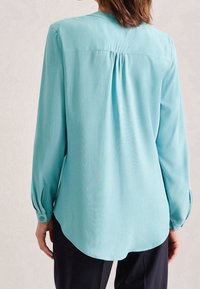 Blusa azzurro chiaro con maniche lunghe, orli arrotondati, polsini con bottone e schiena plissettata. Realizzata in un tessuto liscio e leggero.