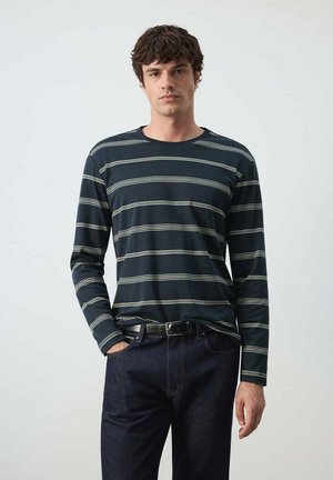 Joven con camisa azul marino de manga larga con rayas horizontales blancas y vaqueros azul oscuro, de pie con una mano en el bolsillo sobre un fondo liso.